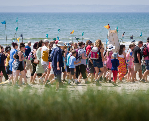Image of a crowd of people of varying ages walking across a beach on a sunny day in Cornwall. They are holding colourful flags.